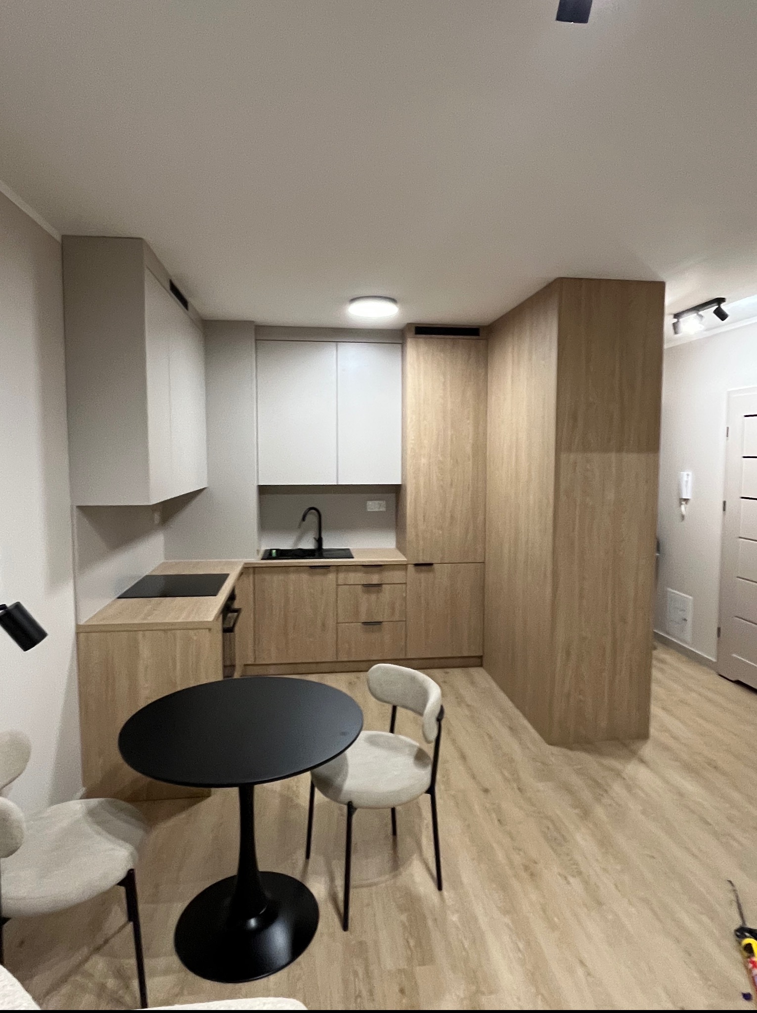 Jasna kuchnia z drewnianymi frontami i białymi szafkami wiszącymi. Na pierwszym planie czarny stół z krzesłami. Nowoczesny design, minimalistyczne wykończenie.