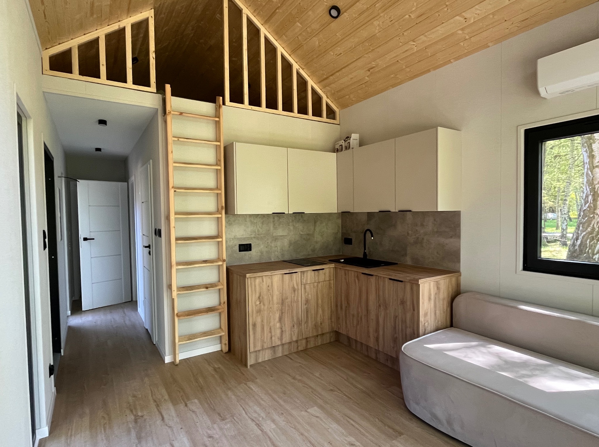 Drewniana drabina prowadzi na antresolę w jasnej kuchni z drewnianymi szafkami i szarą ścianą. W tle widoczny fragment sofy i okno z widokiem na zieleń.