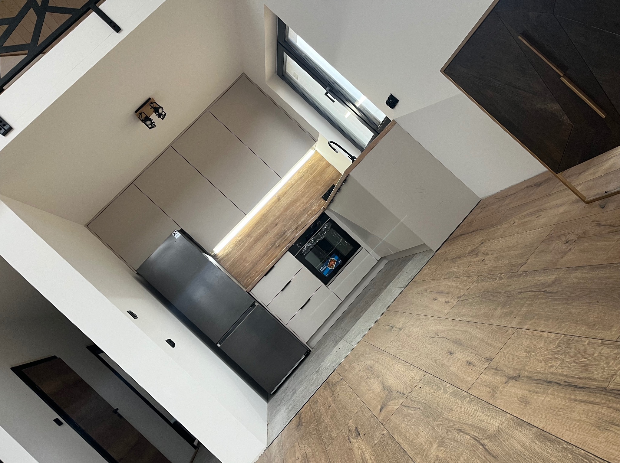 Nowoczesna kuchnia z beżowymi szafkami, drewnianym blatem i czarną lodówką. Ujęcie z góry podkreśla minimalistyczny design i oświetlenie LED pod szafkami.