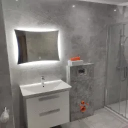 Nowoczesna łazienka w szarościach z podświetlanym lustrem, umywalką z szafką i kabiną prysznicową typu walk-in; widoczne elementy instalacji hydraulicznej.