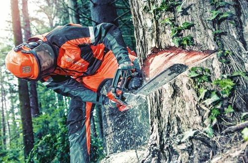 Drwal w pomarańczowym kombinezonie tnący drzewo piłą łańcuchową w lesie, wióry lecą w powietrzu.