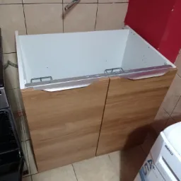 Szafka pod umywalkę z drewnopodobnymi frontami, białe wnętrze, widoczna armatura i pralka, płytki ceramiczne na ścianie i podłodze.