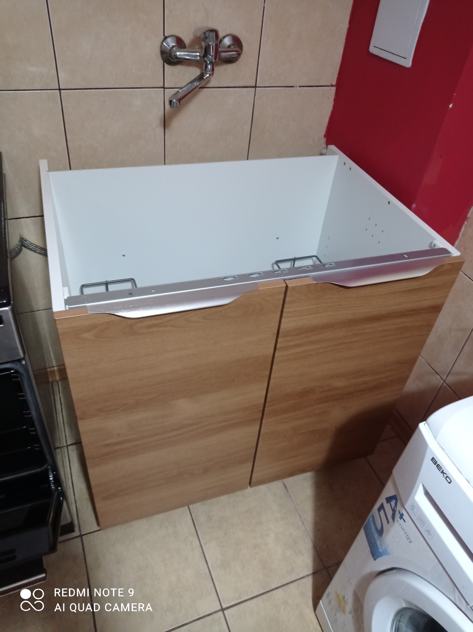 Szafka pod umywalkę z drewnopodobnymi frontami, białe wnętrze, widoczna armatura i pralka, płytki ceramiczne na ścianie i podłodze.