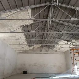 Metalowa konstrukcja dachu hali częściowo pokryta białą pianką izolacyjną, z widocznym rusztowaniem i osobą w kombinezonie ochronnym na nim, w trakcie aplikacji pianki.