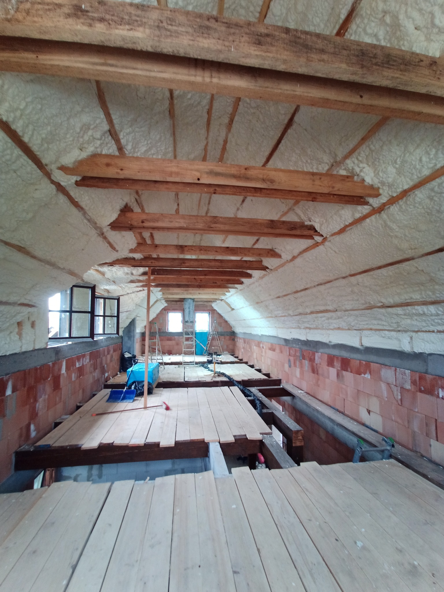 Poddasze w trakcie ocieplania pianką, widoczne drewniane krokwie i biała pianka izolacyjna, częściowo ułożona drewniana podłoga, okna dachowe i drabina w tle.