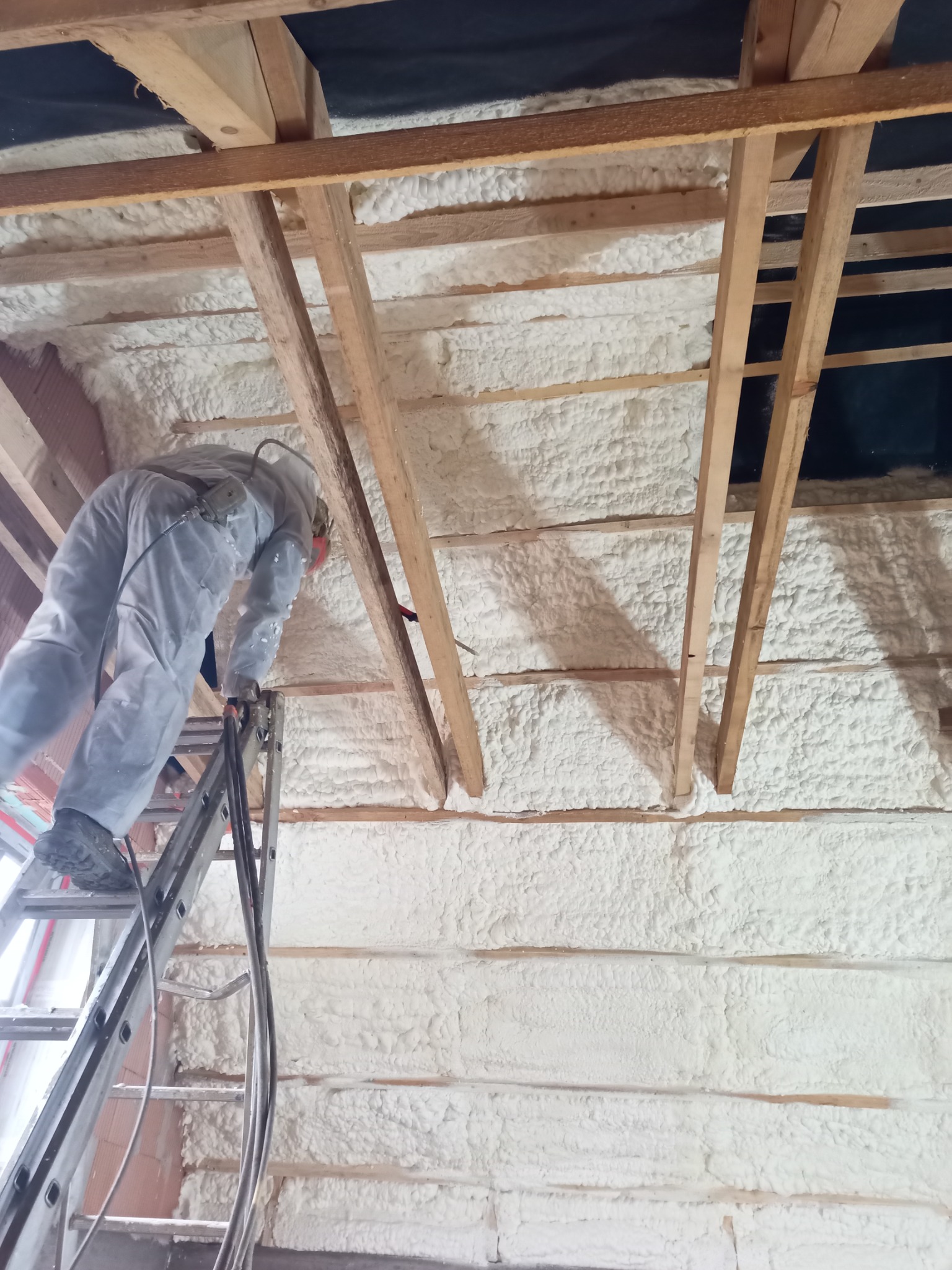 Pracownik w kombinezonie ochronnym na drabinie aplikuje białą piankę izolacyjną na poddasze w Międzyrzeczu Górnym, widoczne drewniane elementy konstrukcji dachu.