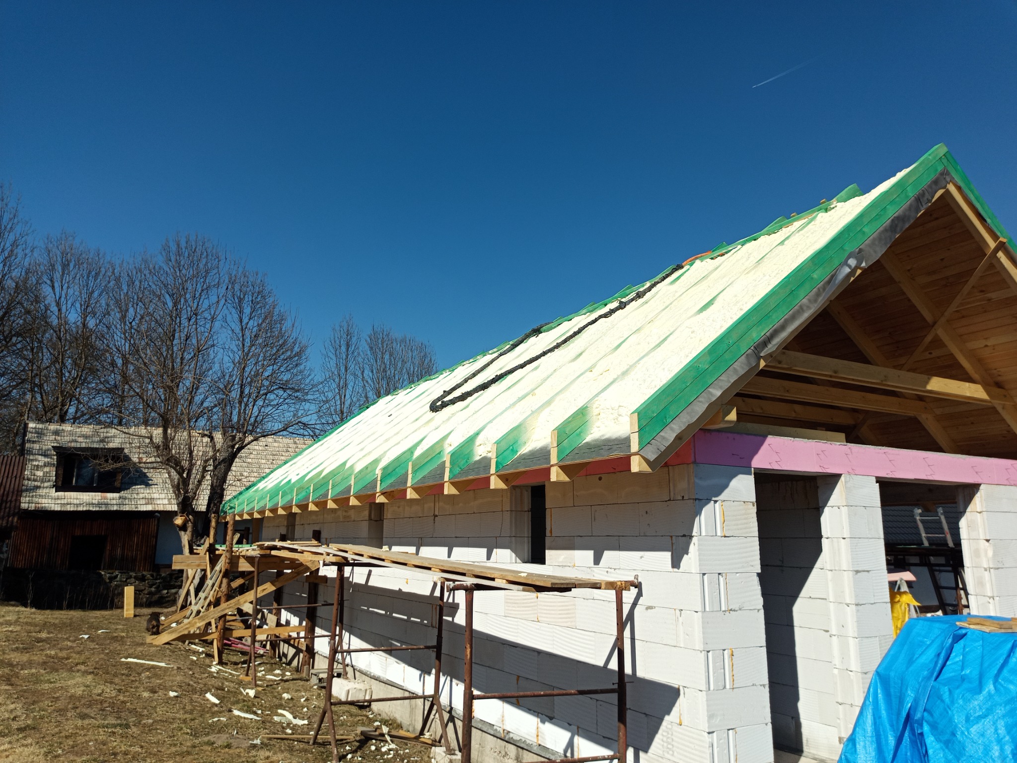Budowa domu w trakcie ocieplania pianką PUR na dachu, widoczne rusztowanie, białe bloczki i niebieska plandeka, w tle stary drewniany dom.