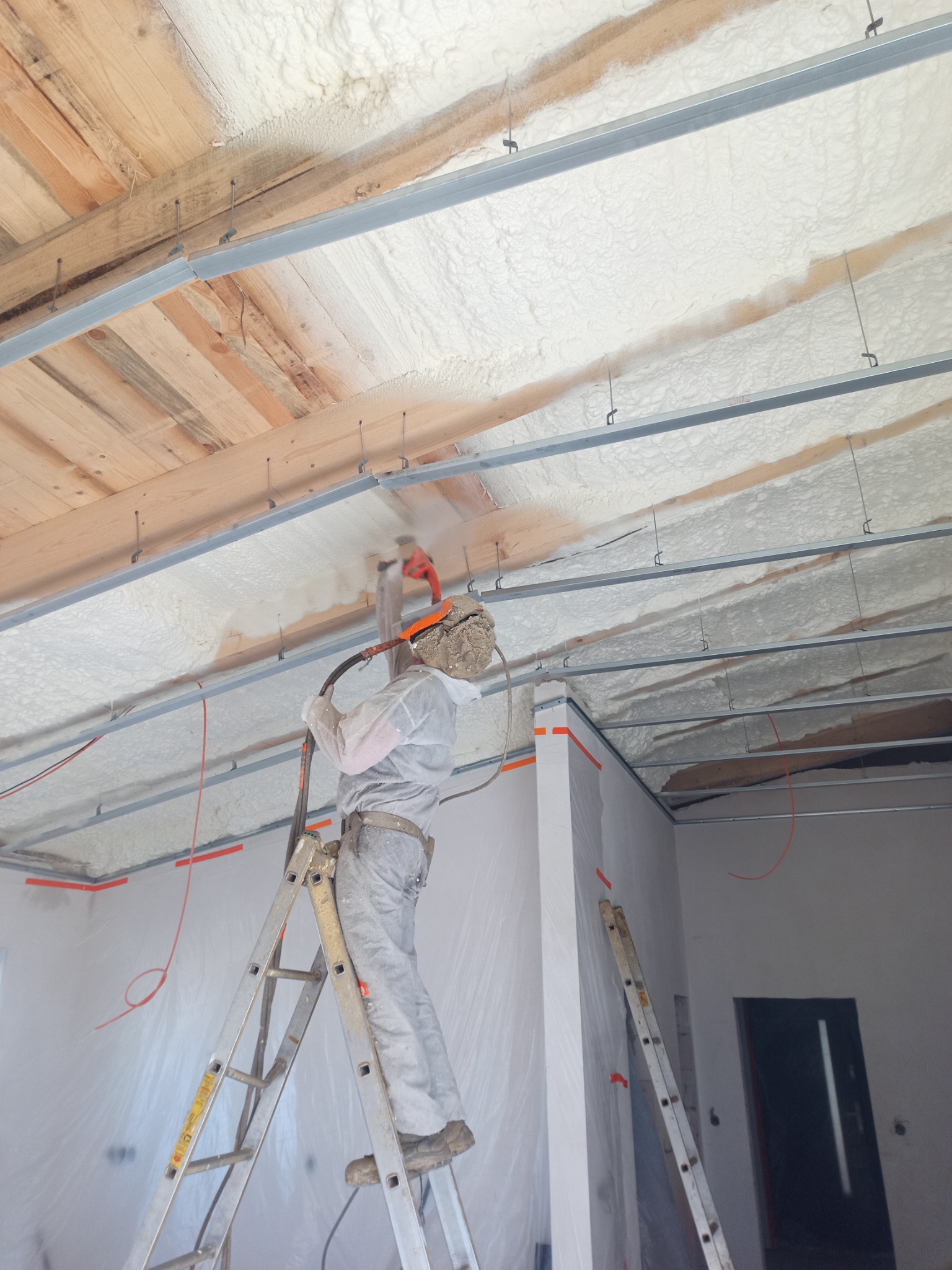 Pracownik w kombinezonie ochronnym na drabinie aplikuje białą pianę izolacyjną na drewniany strop z metalowymi profilami wewnątrz budynku.