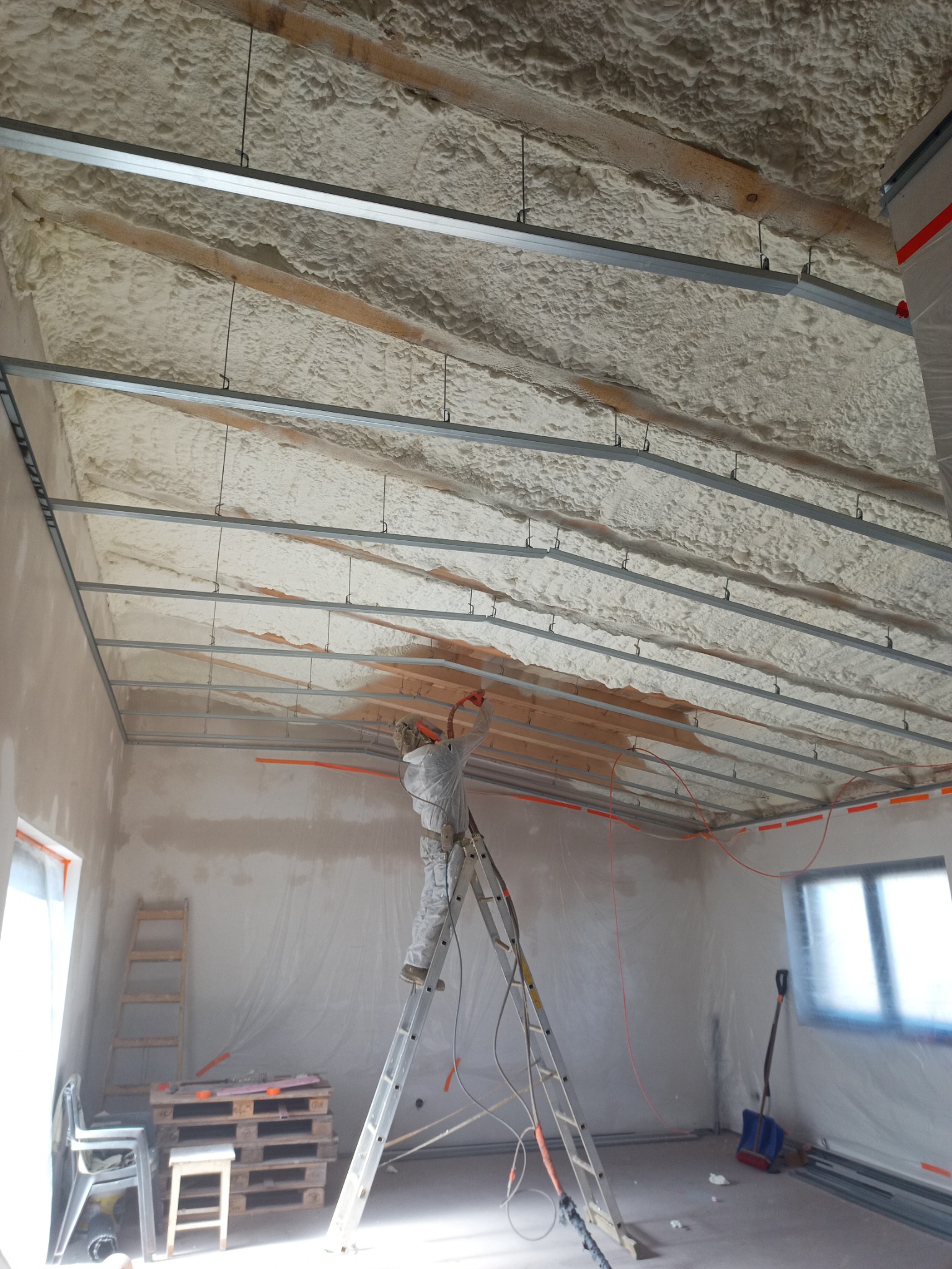 Pracownik w kombinezonie ochronnym na aluminiowej drabinie aplikuje piankę izolacyjną na poddasze, widoczne drewniane belki i metalowe profile konstrukcyjne, pomieszczenie przygotowane folią ochronną.
