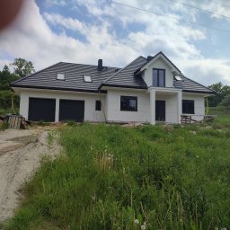 OKUPATO KAMIL WOLNY - Nowoczesny, parterowy dom z dwustanowiskowym garażem, wykończony w bieli z czarnymi akcentami dachu i okien, otoczony bujną zielenią i paletami.