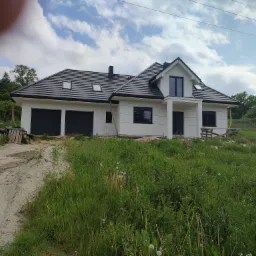 Nowoczesny, parterowy dom z dwustanowiskowym garażem, wykończony w bieli z czarnymi akcentami dachu i okien, otoczony bujną zielenią i paletami.