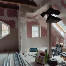 Poddasze w trakcie adaptacji: widoczne płyty karton-gips na skosach i ścianach, szpachlowane łączenia, okna dachowe, narzędzia pracy i profile metalowe na podłodze.