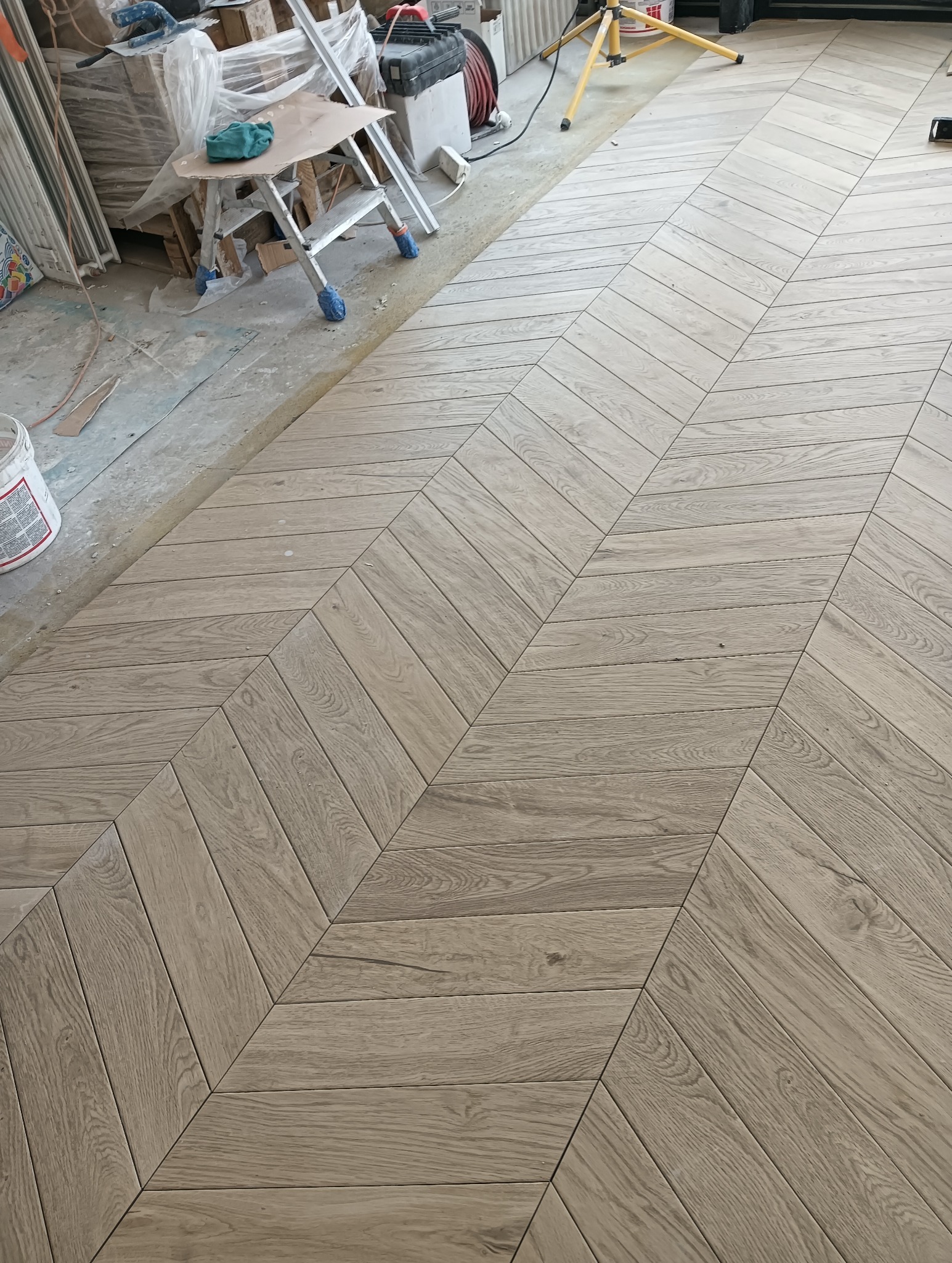 Ułożona parkietowa podłoga w jodełkę w odcieniach szarości i beżu, widok z góry. W tle drabina malarska i materiały budowlane.