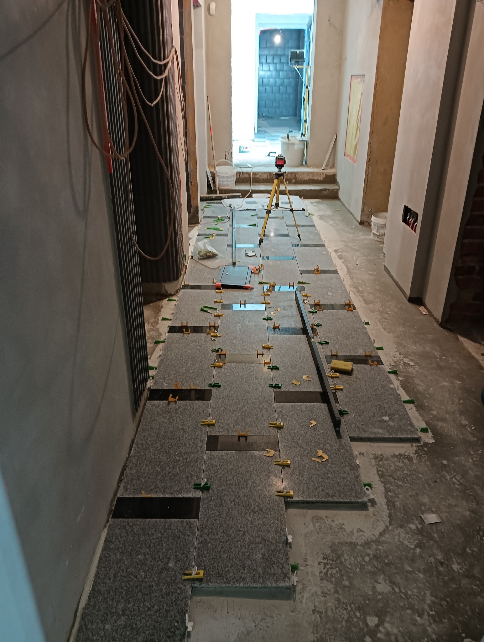 Układanie szarych płytek podłogowych z użyciem poziomicy laserowej i klipsów poziomujących. Widoczny fragment korytarza w trakcie prac wykończeniowych.