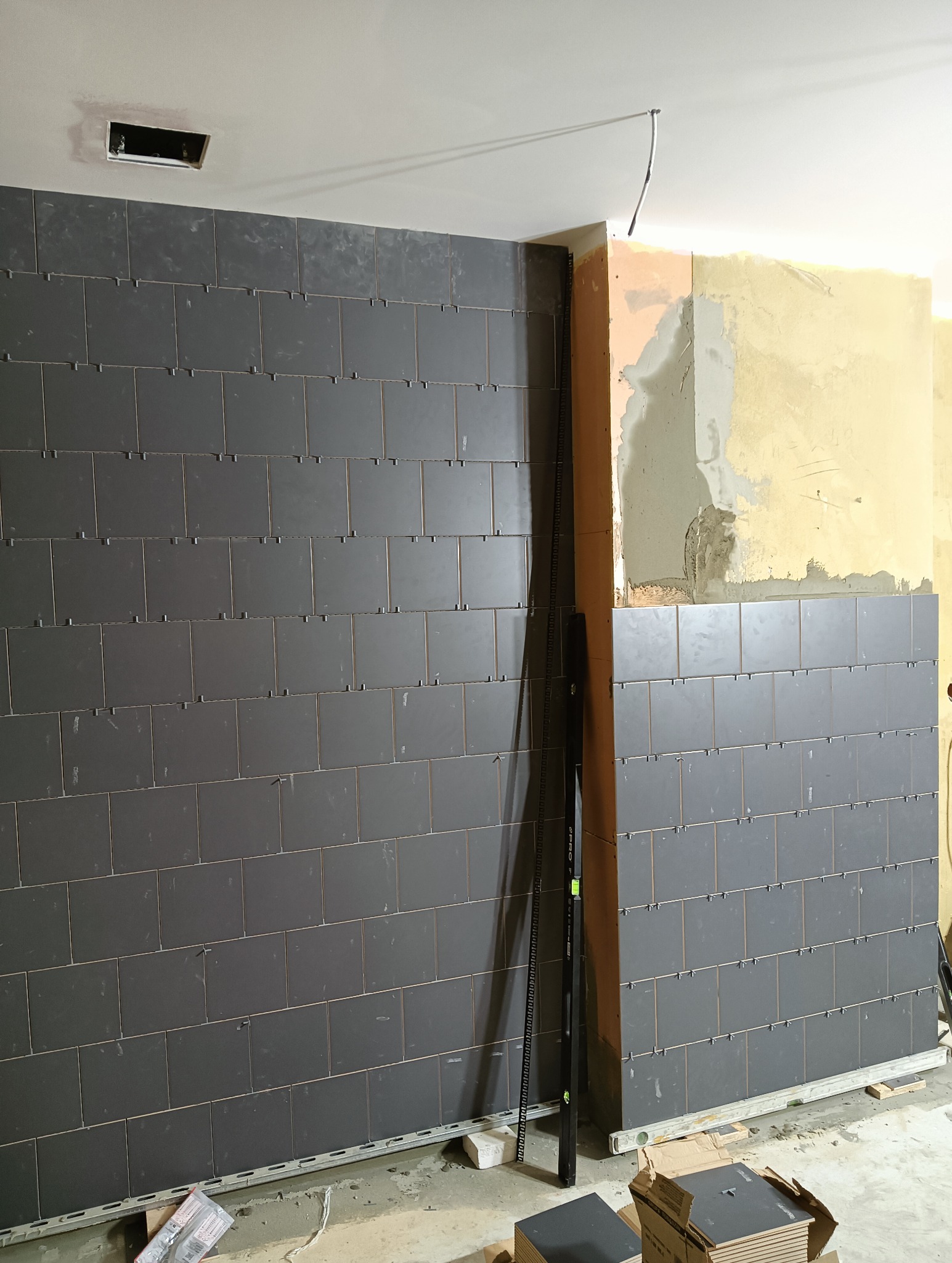 Wnętrze w trakcie wykańczania. Częściowo ułożone ciemne płytki na ścianie, widoczne poziomica i pozostałości po pracach remontowych. Surowy stan pomieszczenia.