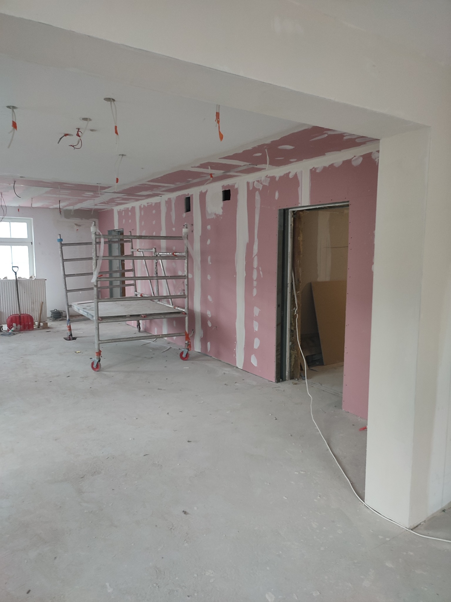 Wnętrze w trakcie prac wykończeniowych: ściany z płyt kartonowo-gipsowych, szpachlowane łączenia, rusztowanie na kółkach i wystające przewody elektryczne.