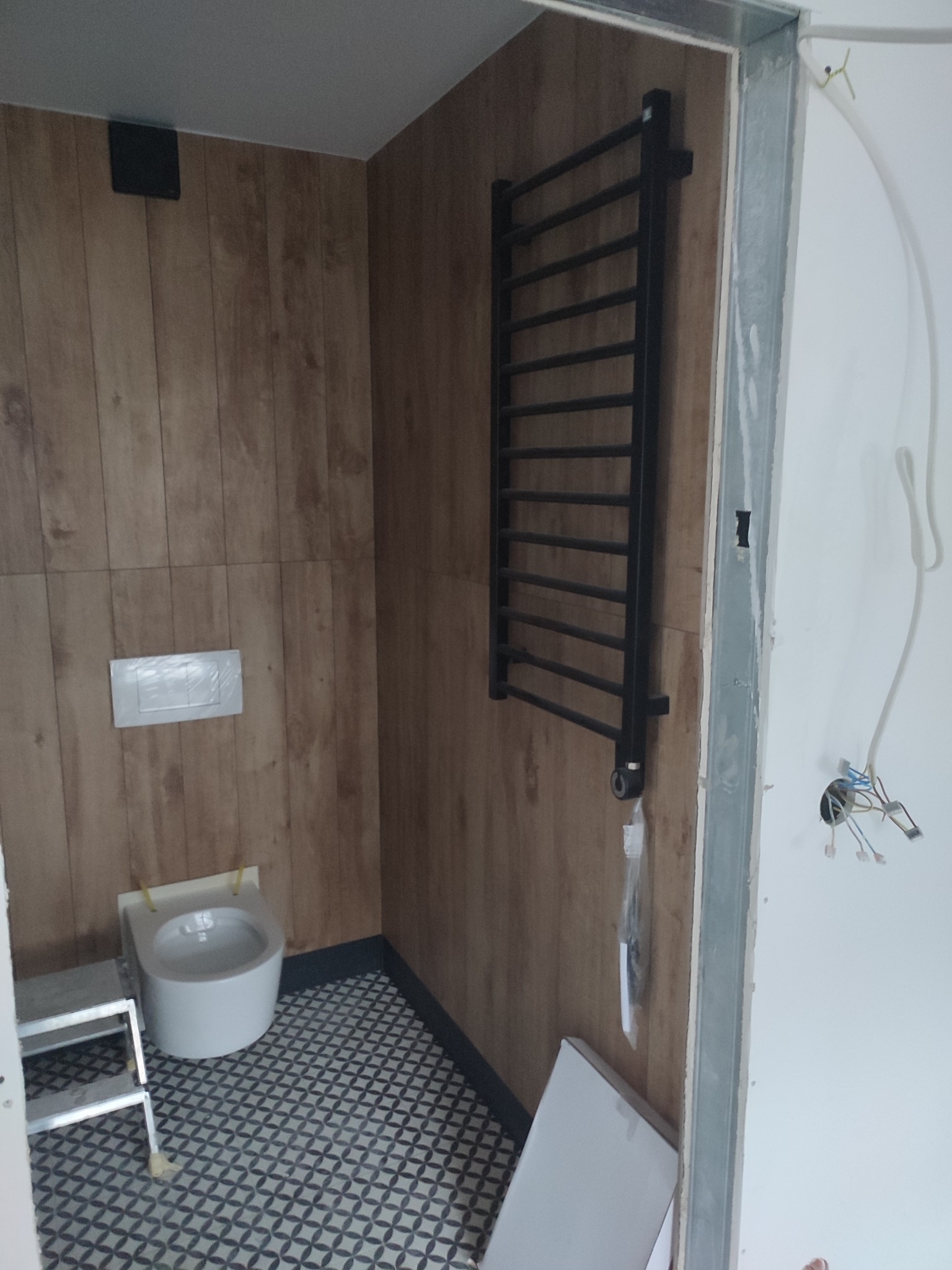 Wnętrze łazienki w trakcie wykończenia: drewnopodobne panele ścienne, wisząca toaleta, grzejnik drabinkowy i fragment instalacji elektrycznej.