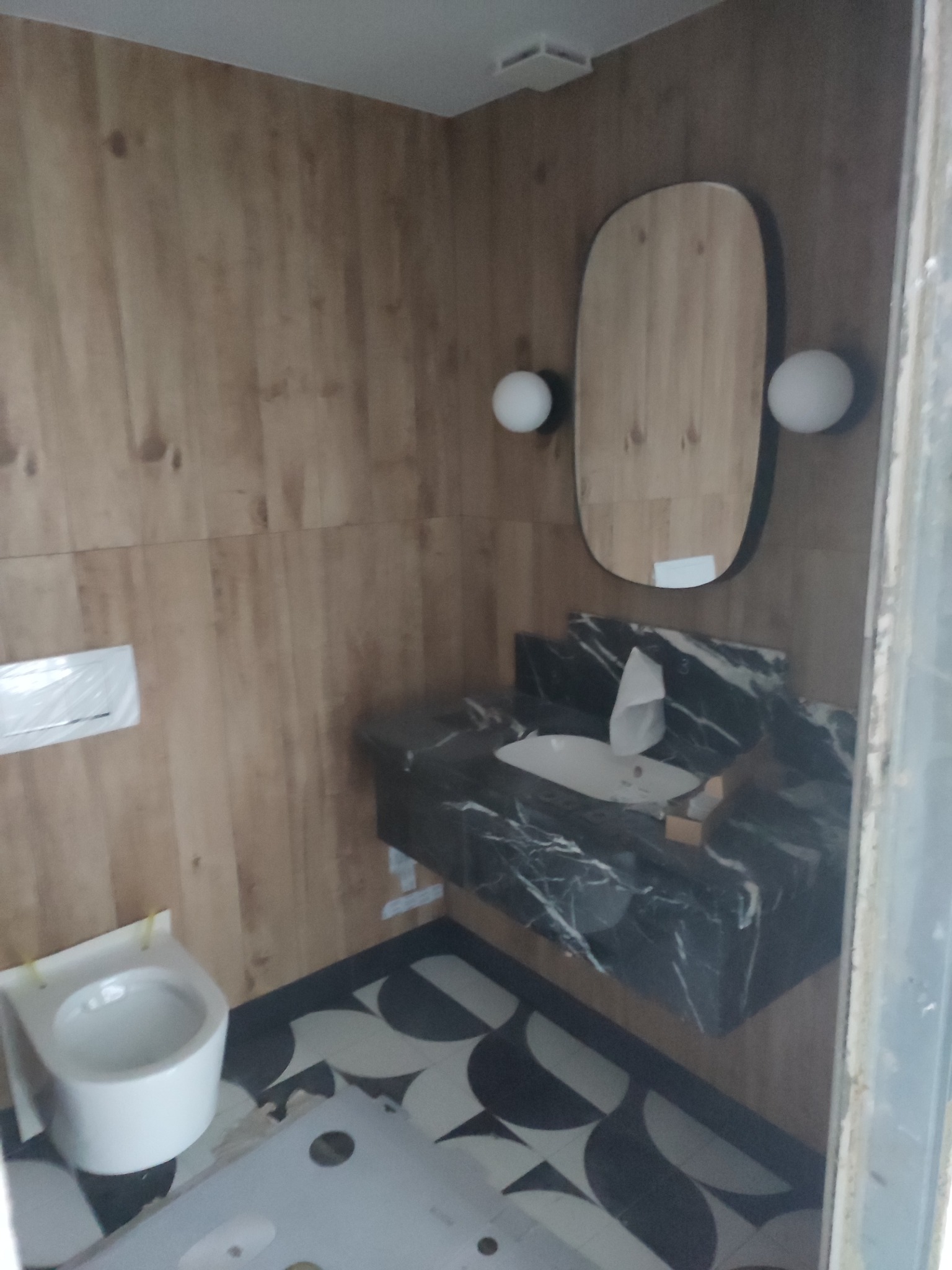 Wykończona łazienka z drewnopodobnymi panelami ściennymi, umywalką z czarnego marmuru i owalnym lustrem z oświetleniem punktowym. Na podłodze wzór w czarno-białe koła.