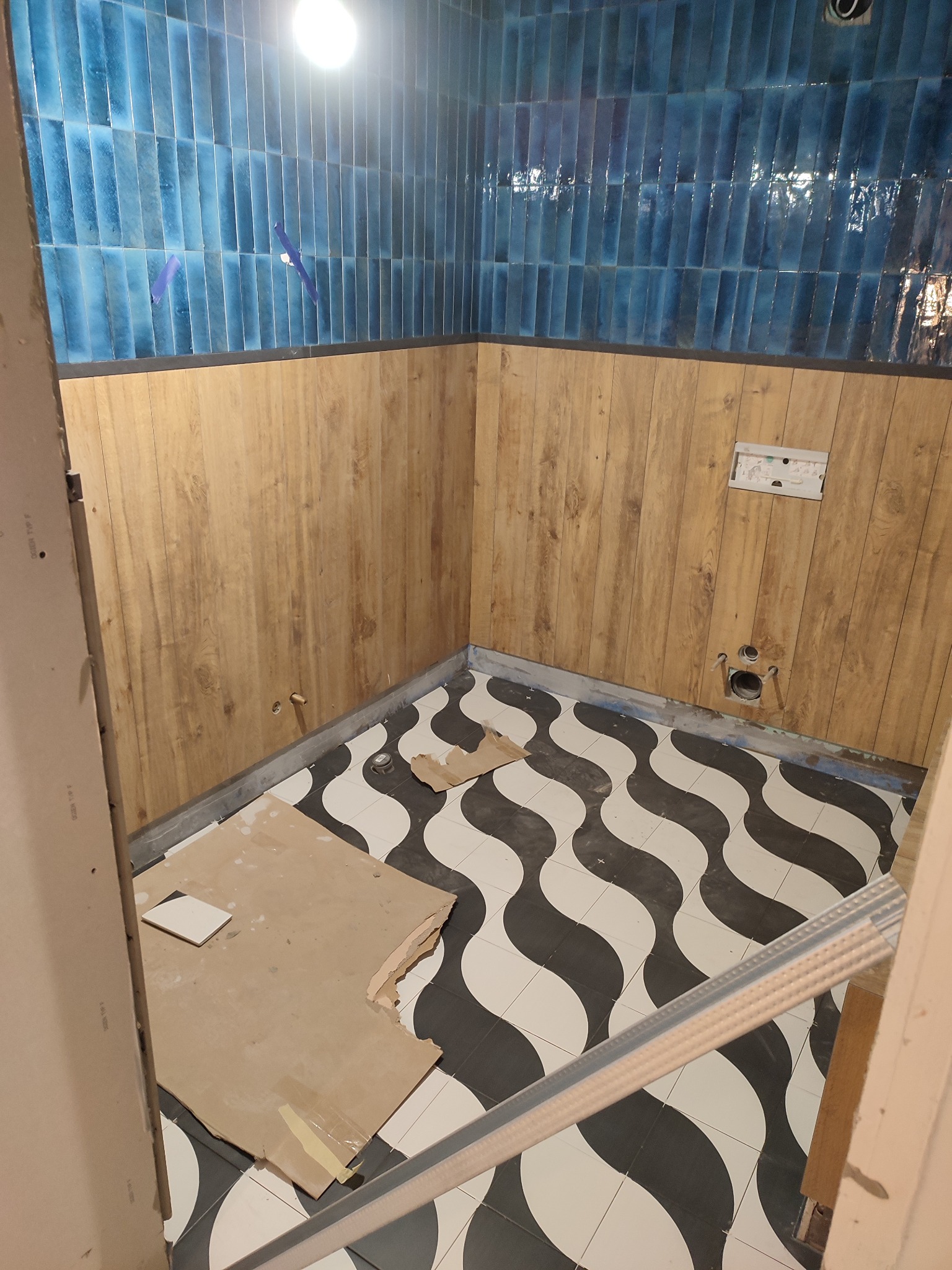 Wnętrze łazienki w trakcie wykończenia: ułożone płytki w odcieniach błękitu na górze, panele imitujące drewno na dole, podłoga w czarno-białe fale. Widoczne elementy instalacji.