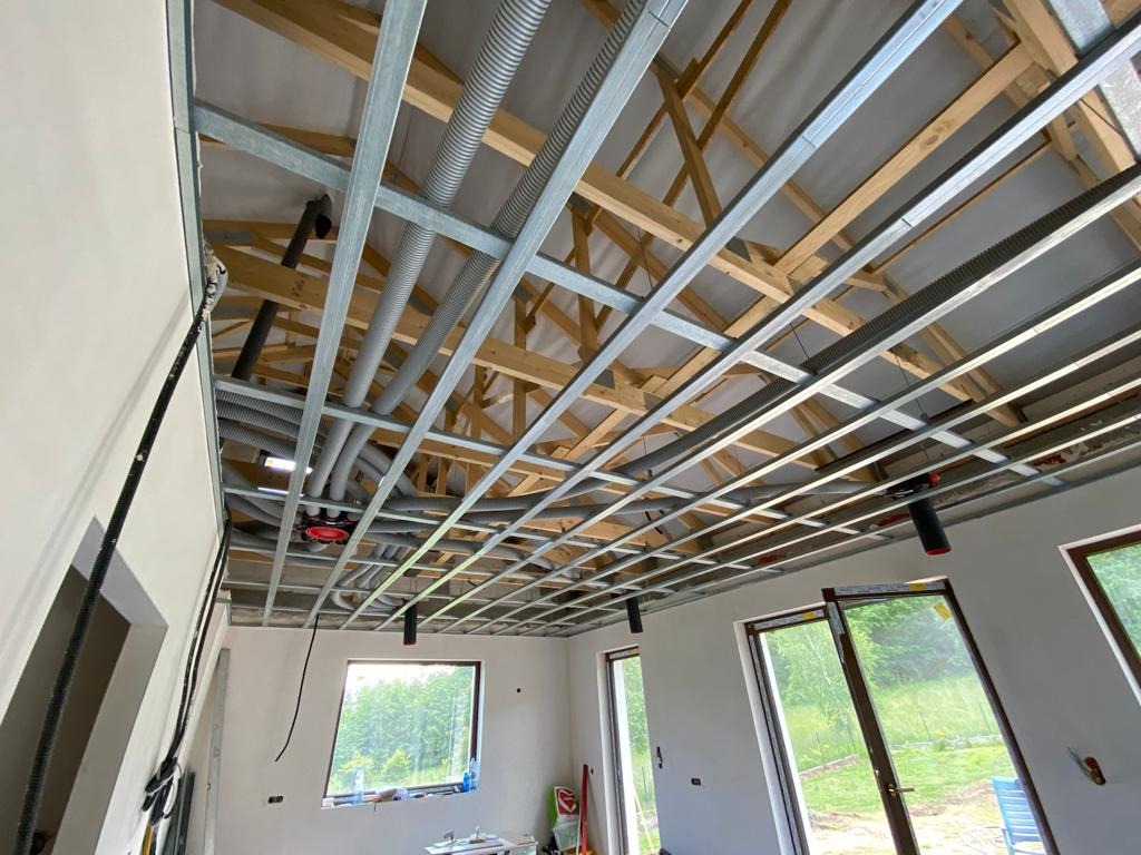 Sufit podwieszany z metalową konstrukcją i instalacją wentylacyjną w surowym wnętrzu domu z oknami. Widoczne drewniane elementy więźby dachowej.