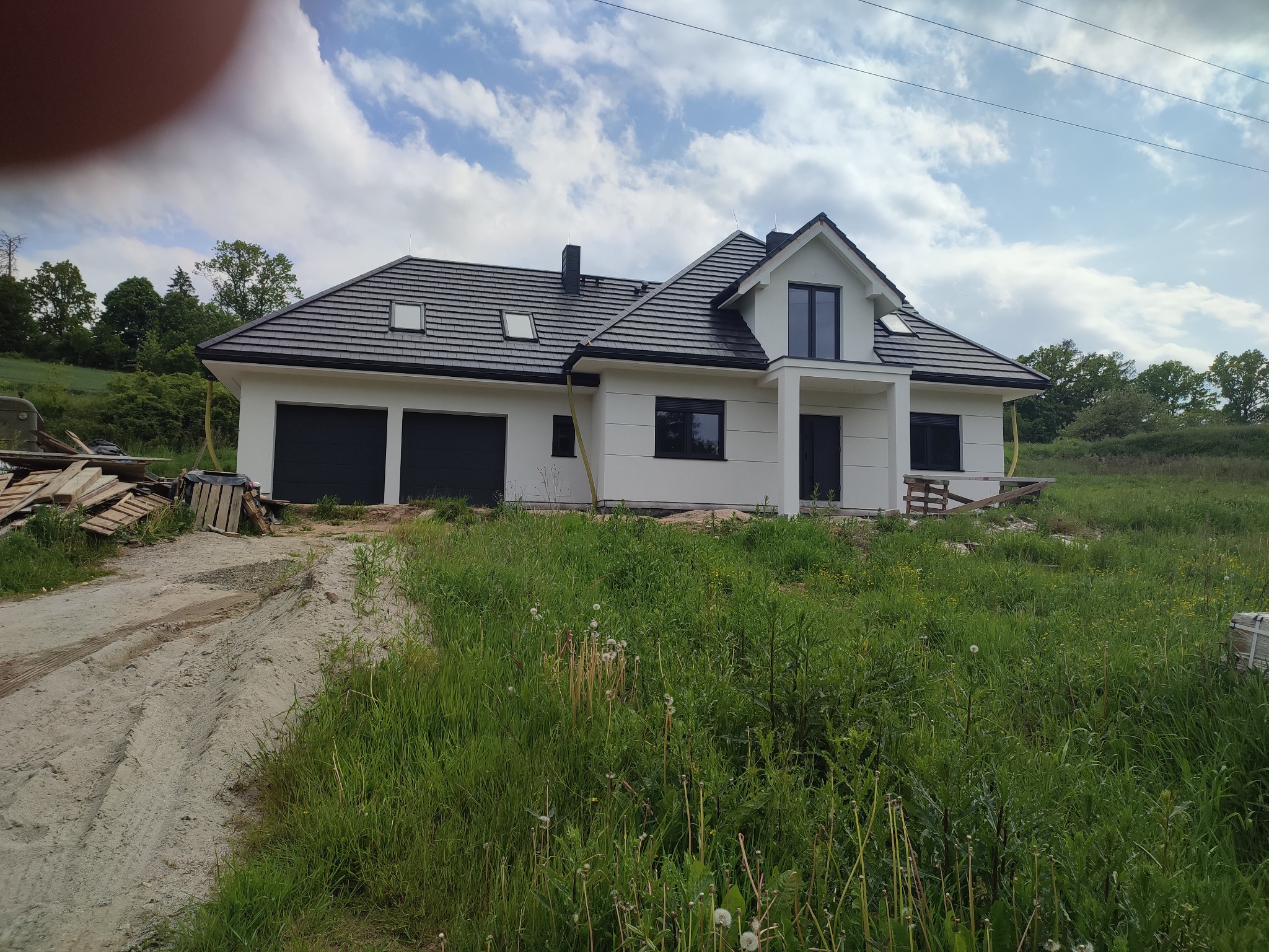 Nowoczesny, parterowy dom z dwustanowiskowym garażem, wykończony w bieli z czarnymi akcentami dachu i okien, otoczony bujną zielenią i paletami.