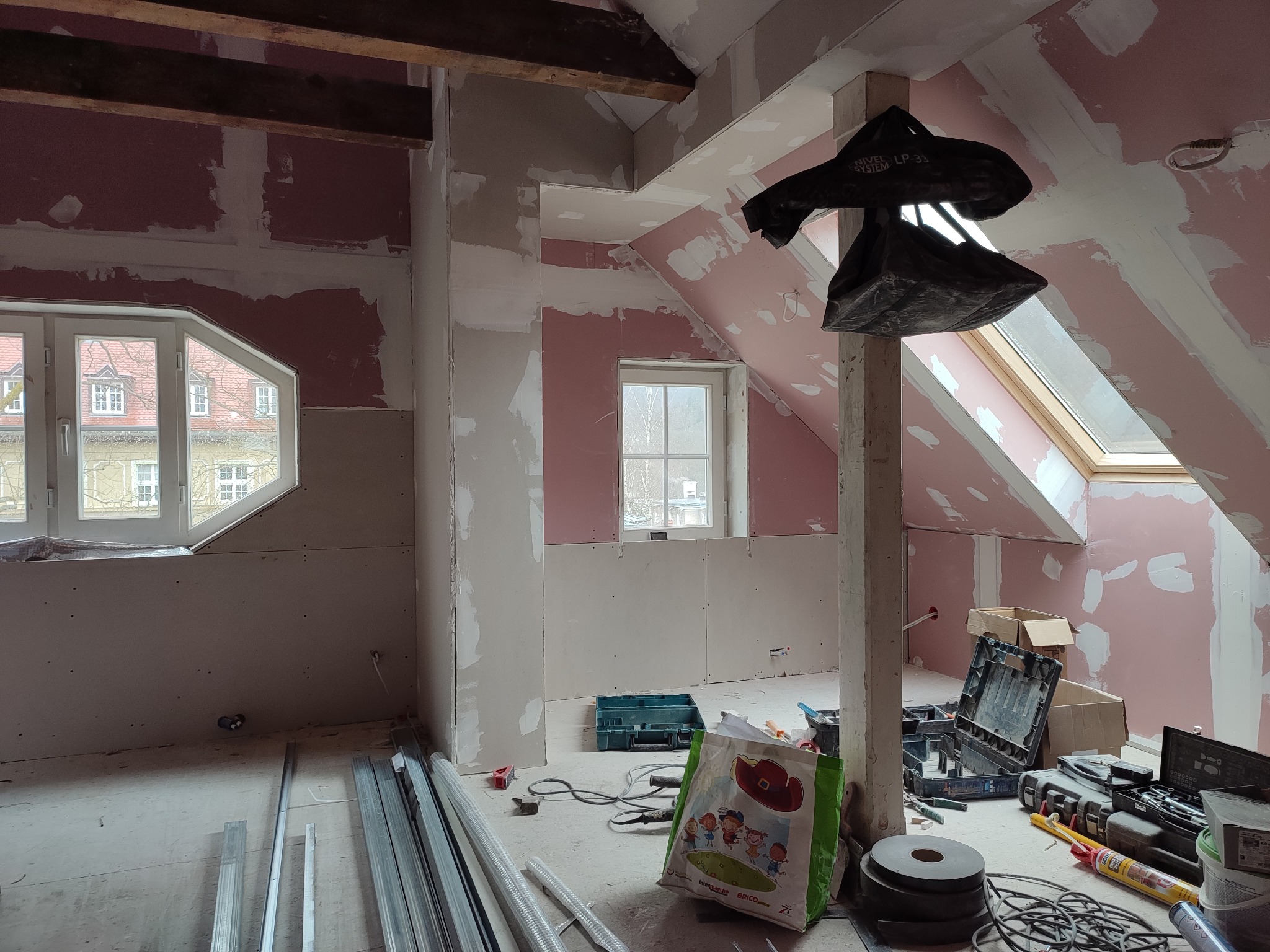 Poddasze w trakcie adaptacji: widoczne płyty karton-gips na skosach i ścianach, szpachlowane łączenia, okna dachowe, narzędzia pracy i profile metalowe na podłodze.