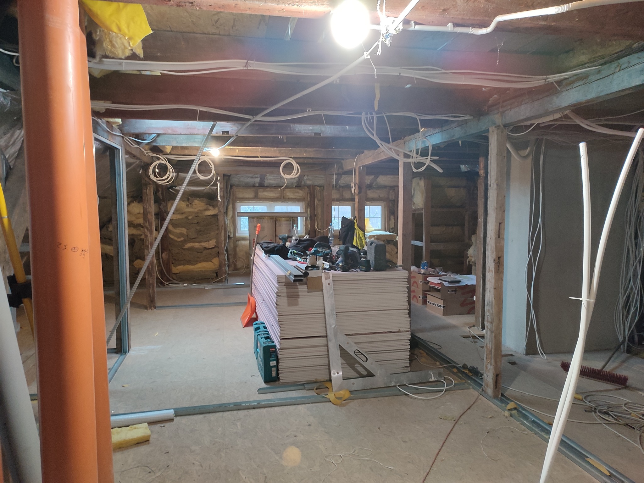 Wnętrze w trakcie prac z płytami gipsowo-kartonowymi: widoczne profile, instalacje elektryczne i stos płyt. Oświetlenie robocze i narzędzia w przestrzeni remontowanej.