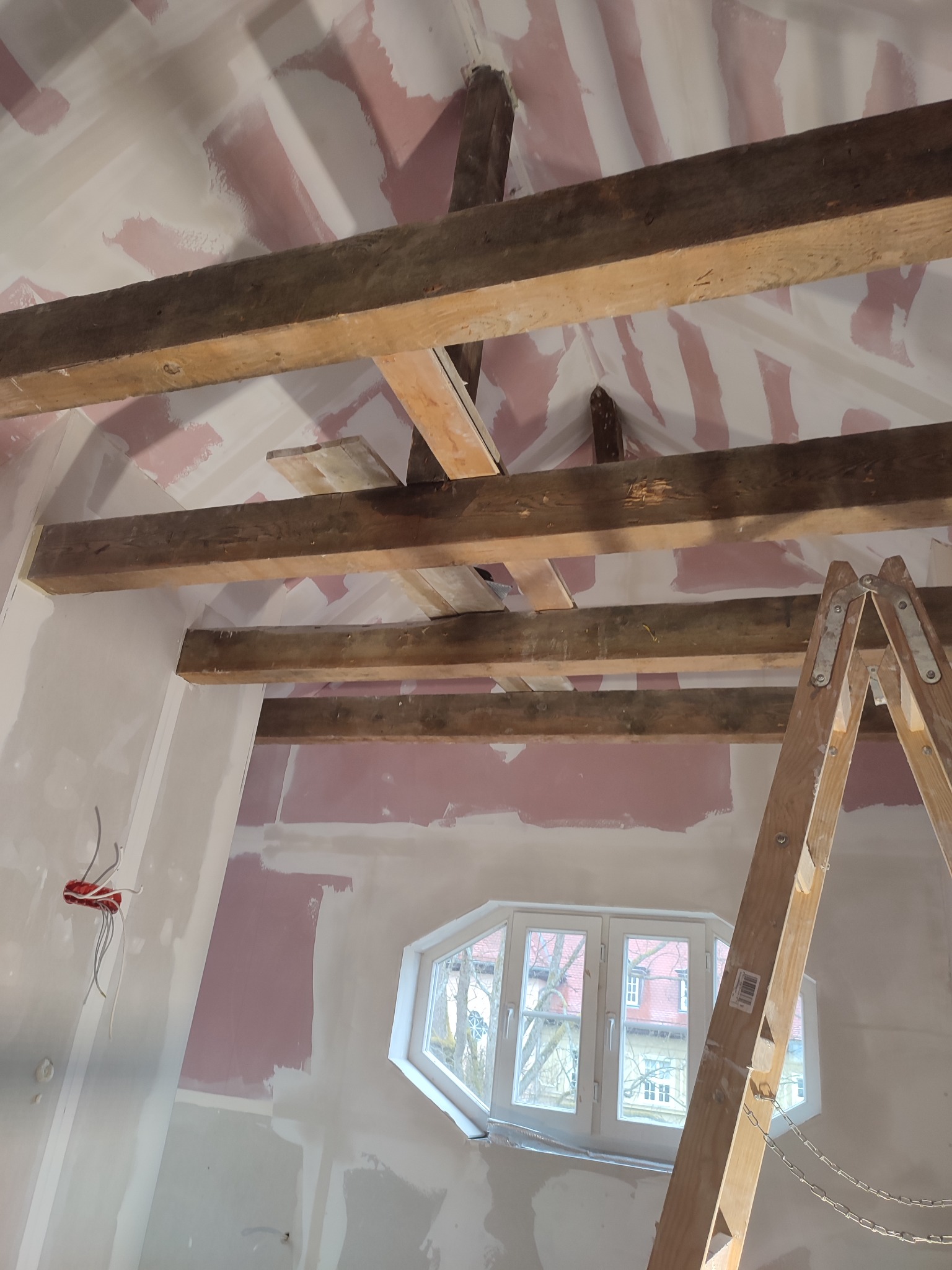 Poddasze w trakcie wykańczania: drewniane belki, ściany z płyt gipsowo-kartonowych, okno w kształcie trapezu, drabina. Widoczne ślady szpachlowania i przygotowania do malowania.