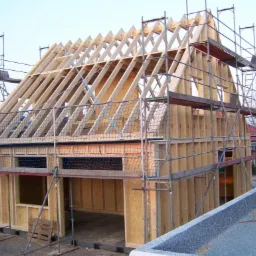 Konstrukcja szkieletowa domu w trakcie budowy, widoczne drewniane elementy ścian i dachu, rusztowania i siatka zabezpieczająca, surowy stan.