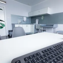 Gabinet lekarski z biurkiem i klawiaturą na pierwszym planie, w tle umywalka, szafki i tablica Snellena do badania wzroku.