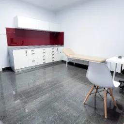 Jasne wnętrze gabinetu lekarskiego z beżowym leżanką, biurkiem, krzesłami i szafkami z czerwonym panelem na ścianie.