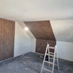 Pokój na poddaszu w Elblągu w trakcie prac wykończeniowych: częściowo położona tapeta o wzorze drewna, drabina, widoczne narzędzia i akcesoria malarskie.