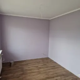 Pomalowane na liliowo wnętrze pokoju z panelami podłogowymi, widoczny grzejnik, puszka po farbie i wystający kabel z sufitu.