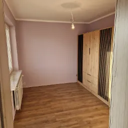 Pusty pokój po remoncie z drewnianą podłogą, świeżo pomalowanymi ścianami w odcieniu liliowym i nowoczesną szafą z drewnianymi i czarnymi listwami, widok zza framugi drzwi.