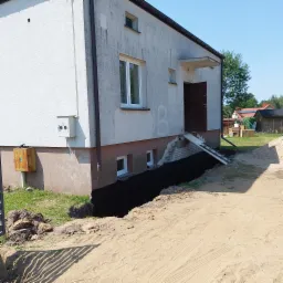 Wykop fundamentów wokół domu z elewacją w trakcie przygotowania do ocieplenia, widoczny fragment ceglanej ściany i drabina.