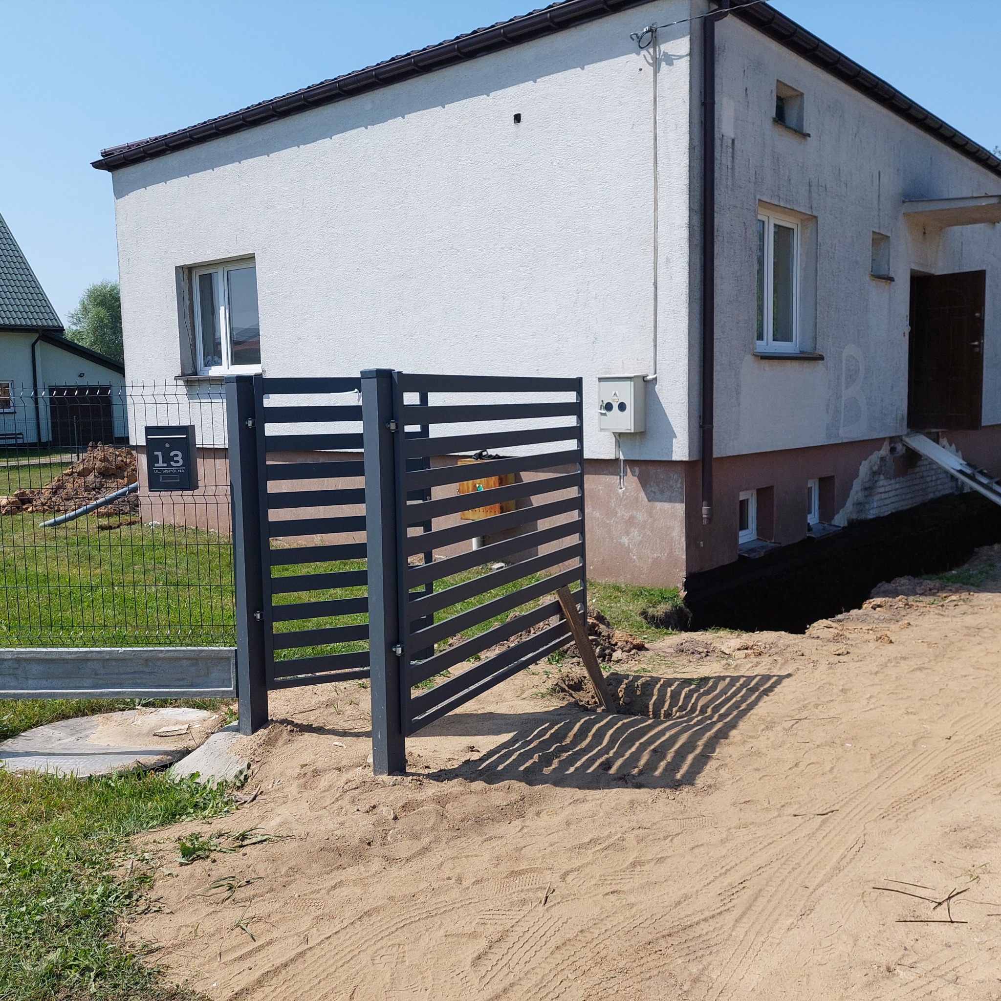 Nowoczesne, poziome przęsła ogrodzeniowe w kolorze grafitowym, częściowo zamontowane przed domem z elewacją w jasnym kolorze, widoczne ślady prac ziemnych.