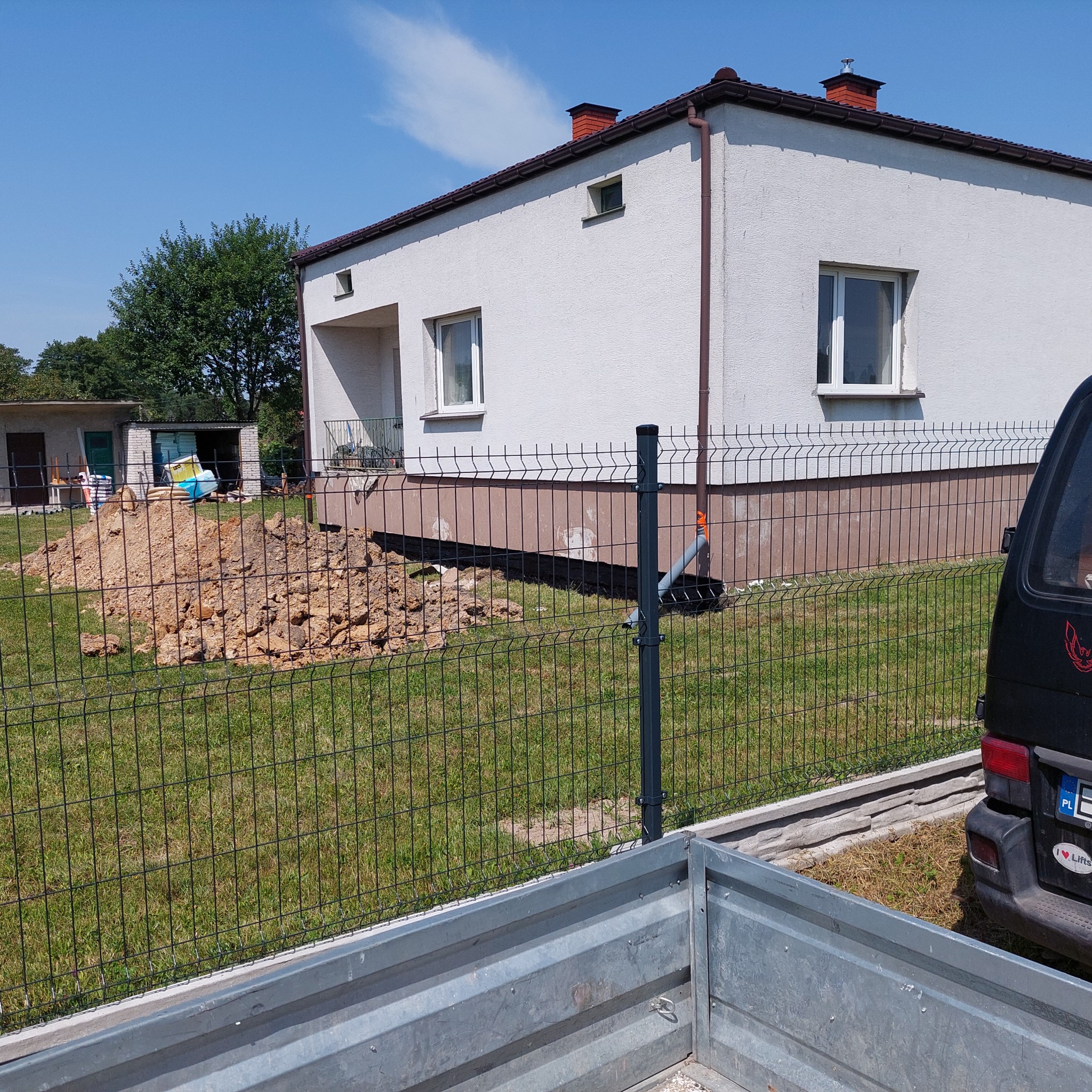 Dwukondygnacyjny dom z białą elewacją i brązowymi akcentami, widoczny zza nowego ogrodzenia panelowego, z wykopaną ziemią obok fundamentów i przyczepą w pierwszym planie.