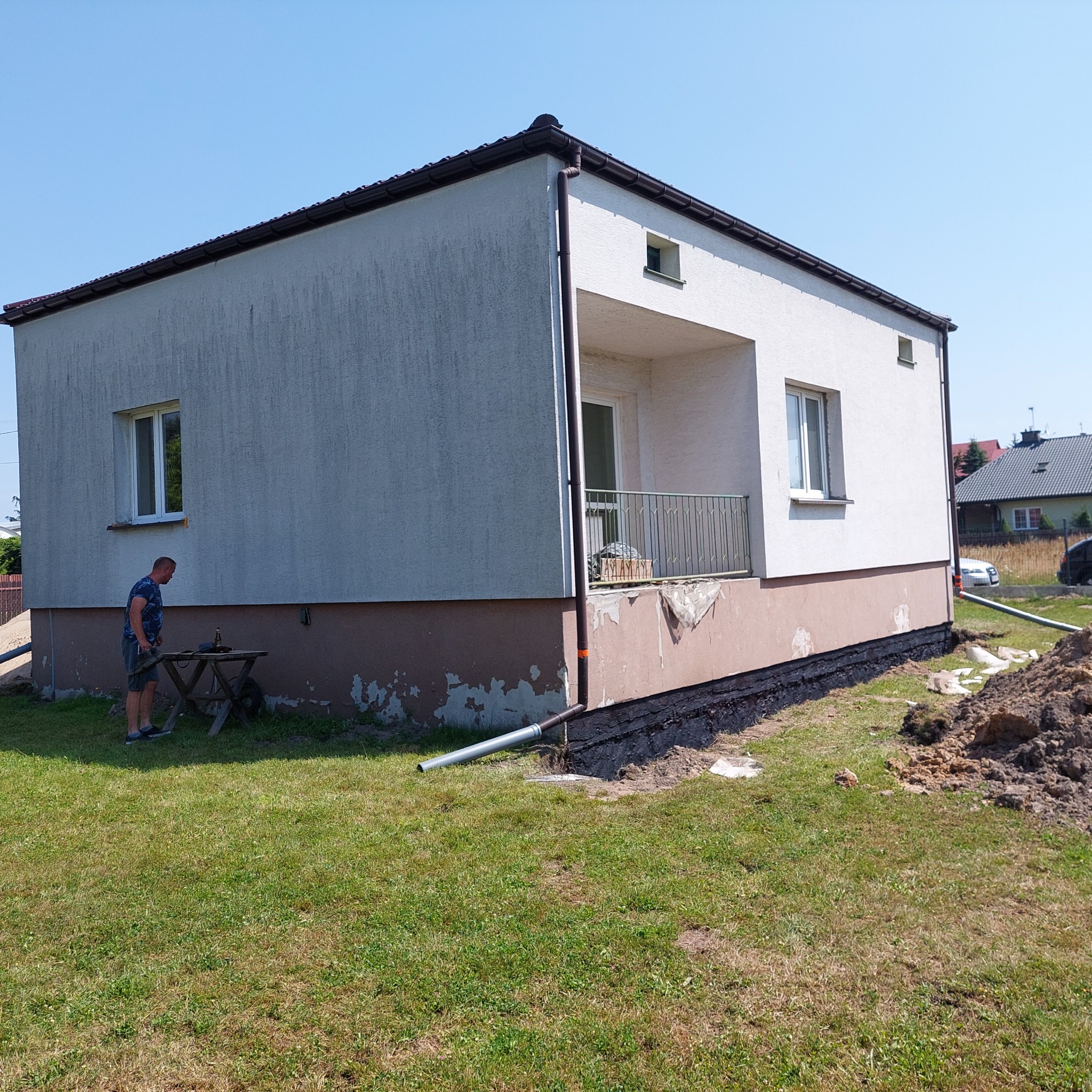Dom jednorodzinny z widoczną izolacją fundamentów w trakcie prac, z fragmentami starej elewacji i przygotowanym terenem, mężczyzna przy stoliku na zewnątrz.