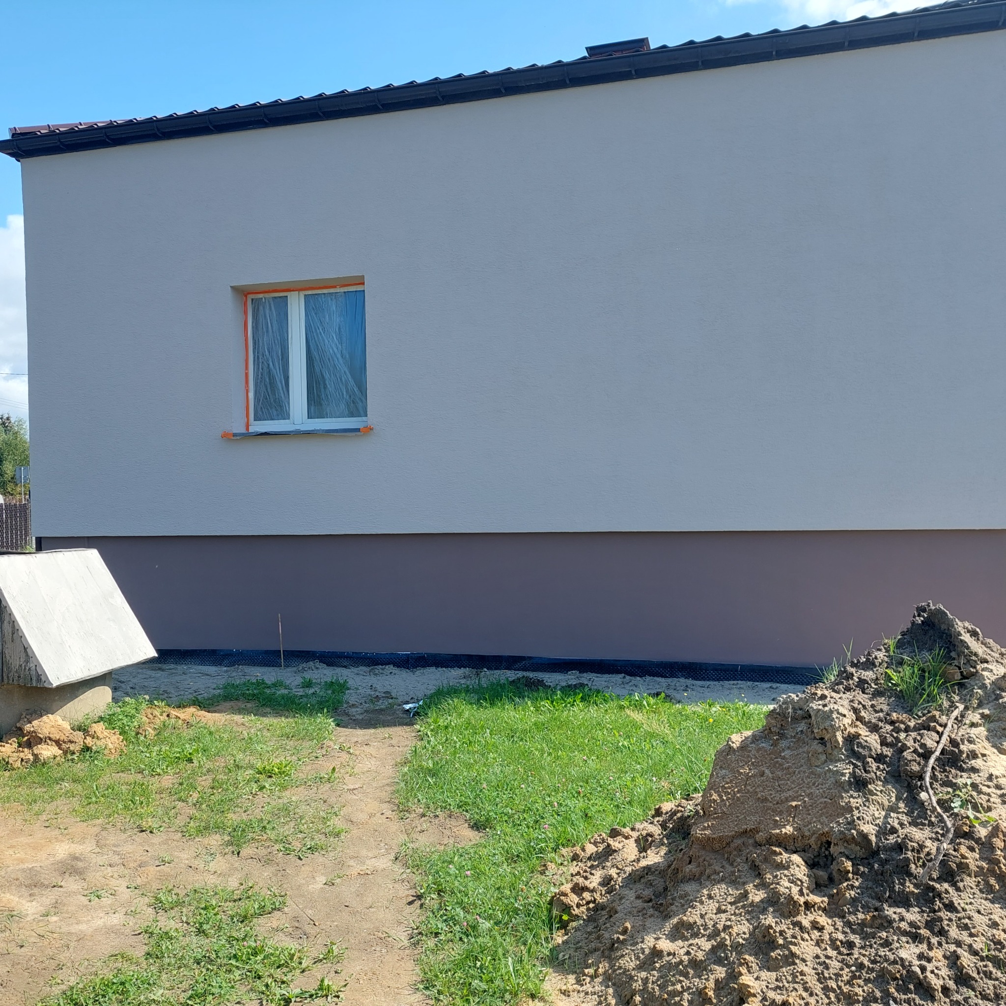 Świeżo otynkowana elewacja domu jednorodzinnego z zabezpieczonym oknem folią malarską, widoczny cokół w kolorze brązowym i sterta ziemi na trawniku.