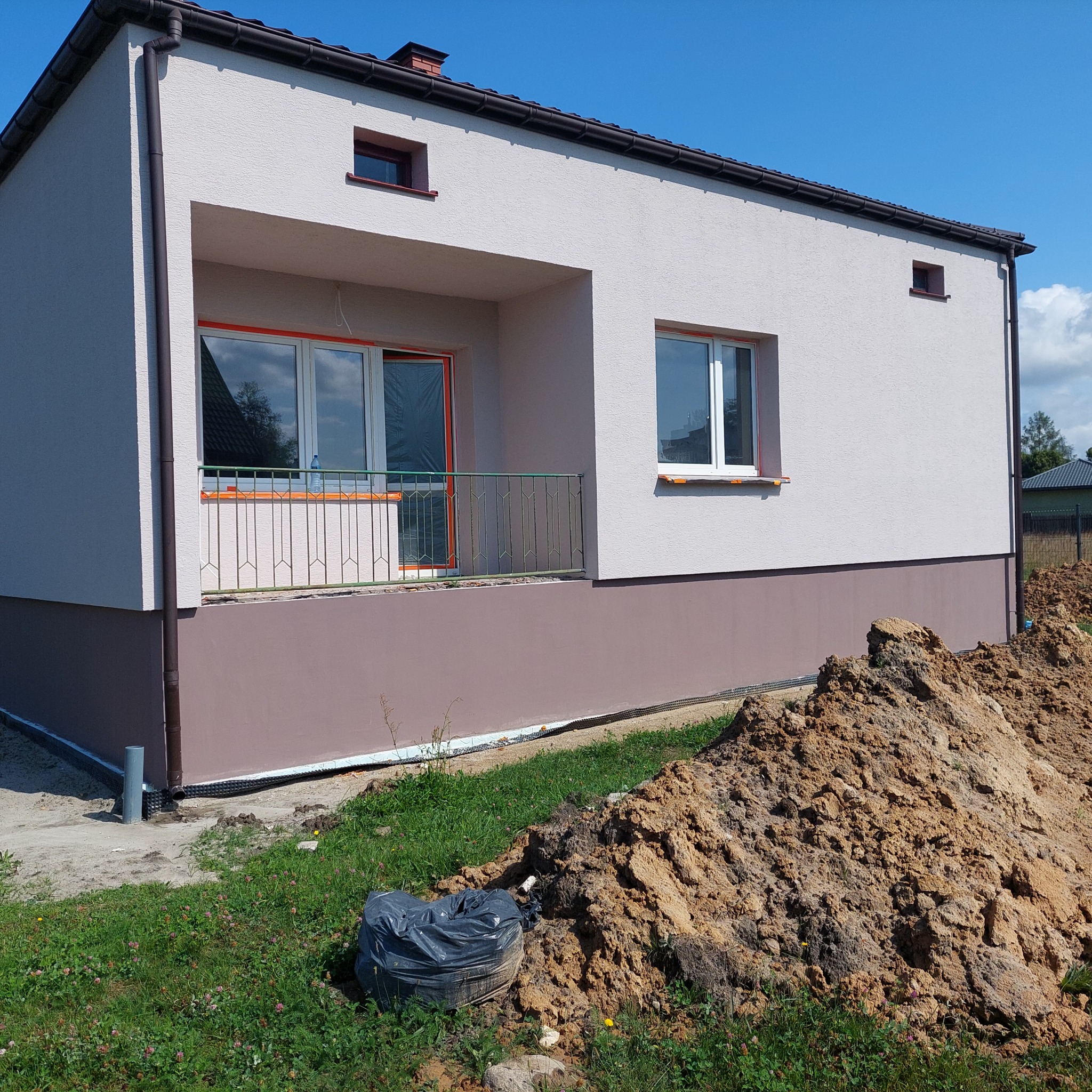 Nowo wybudowany dom jednorodzinny z dwukolorową elewacją, widoczne okna zabezpieczone folią ochronną, obok sterta ziemi i czarny worek na śmieci.