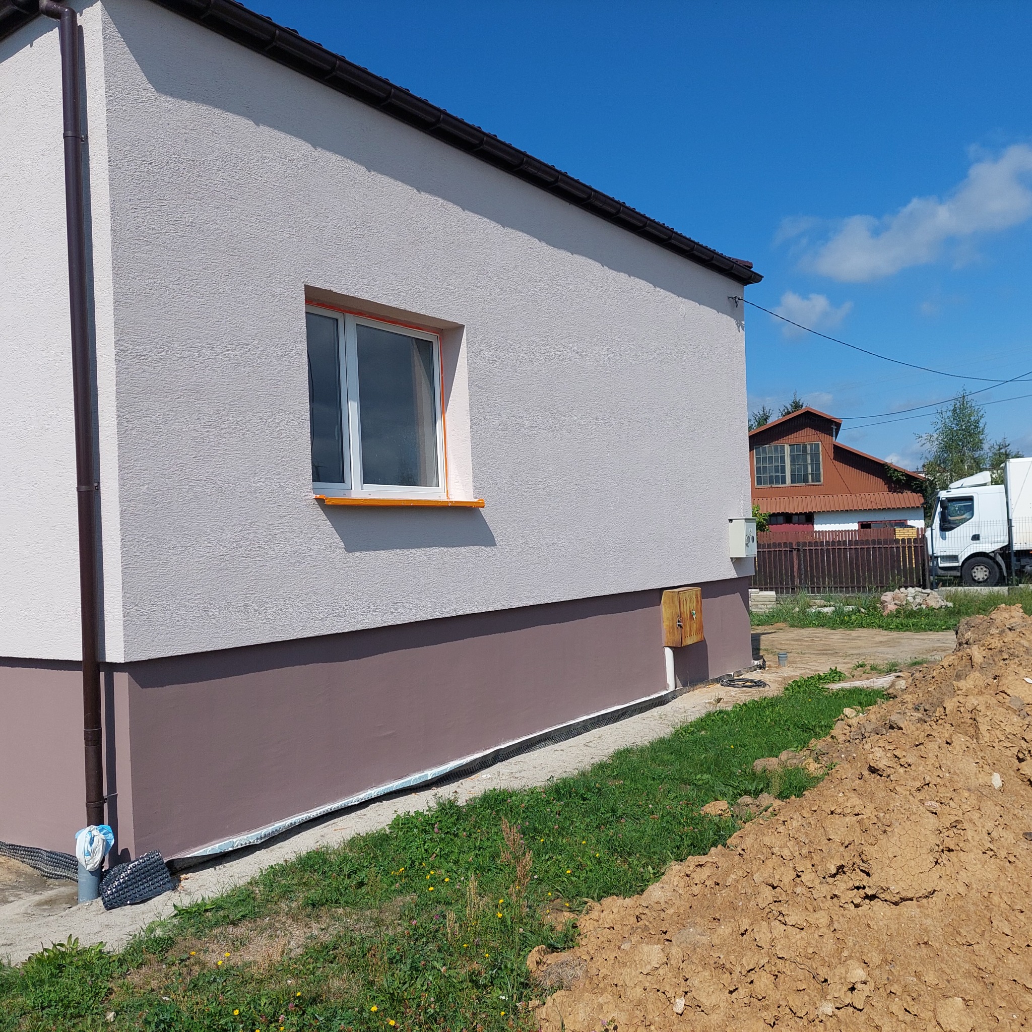 Wykończona elewacja domu jednorodzinnego z jasnoszarym tynkiem strukturalnym i brązowym cokołem, okno zabezpieczone pomarańczową taśmą malarską, widoczny fragment podwórka z ziemią.