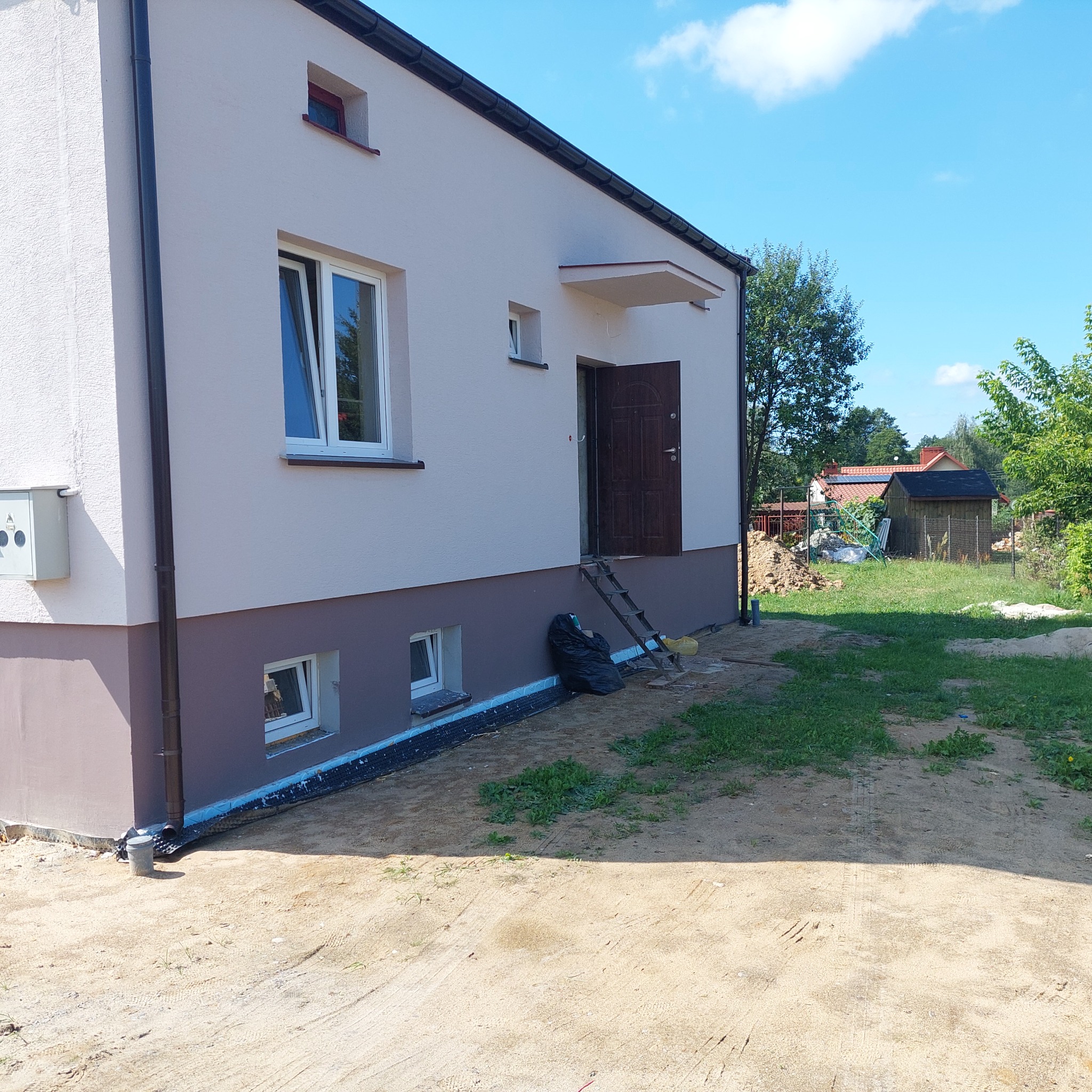 Dwukondygnacyjny dom z nową, jasną elewacją i ciemnobrązowym cokołem, widoczne okna z białymi ramami, rynna, schodki przy wejściu i czarny worek ze śmieciami obok, teren wokół domu w trakcie prac...