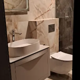 Wykończona łazienka z beżowymi płytkami imitującymi marmur na ścianach i podłodze, owalna umywalka nablatowa na szarej szafce, wisząca toaleta i szklana kabina prysznicowa z ciemnymi płytkami.