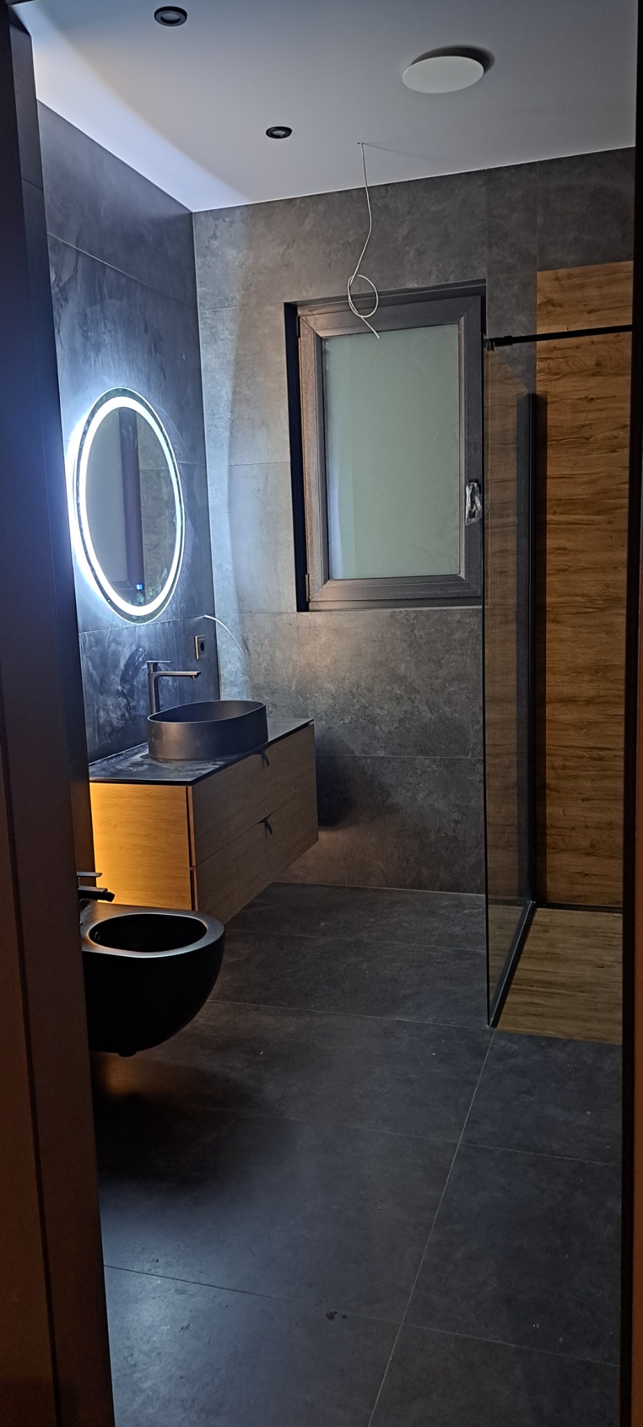 Nowoczesna łazienka z podwieszaną miską WC, umywalką na szafce z drewnianym frontem i okrągłym lustrem z podświetleniem LED na tle szarych płytek imitujących beton.