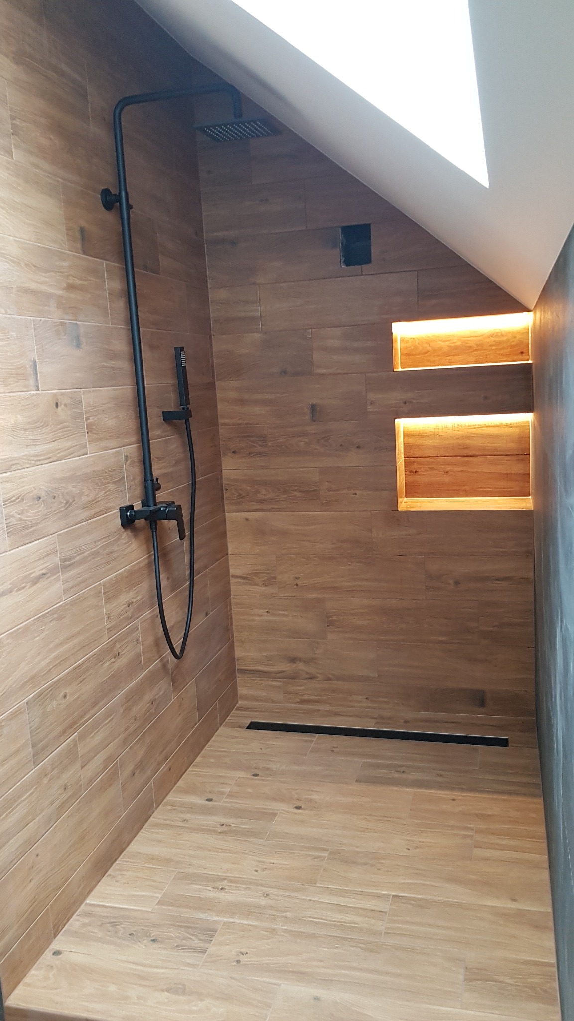Kabina prysznicowa w skosie z drewnopodobnymi płytkami, czarną armaturą i oświetlonymi wnękami.