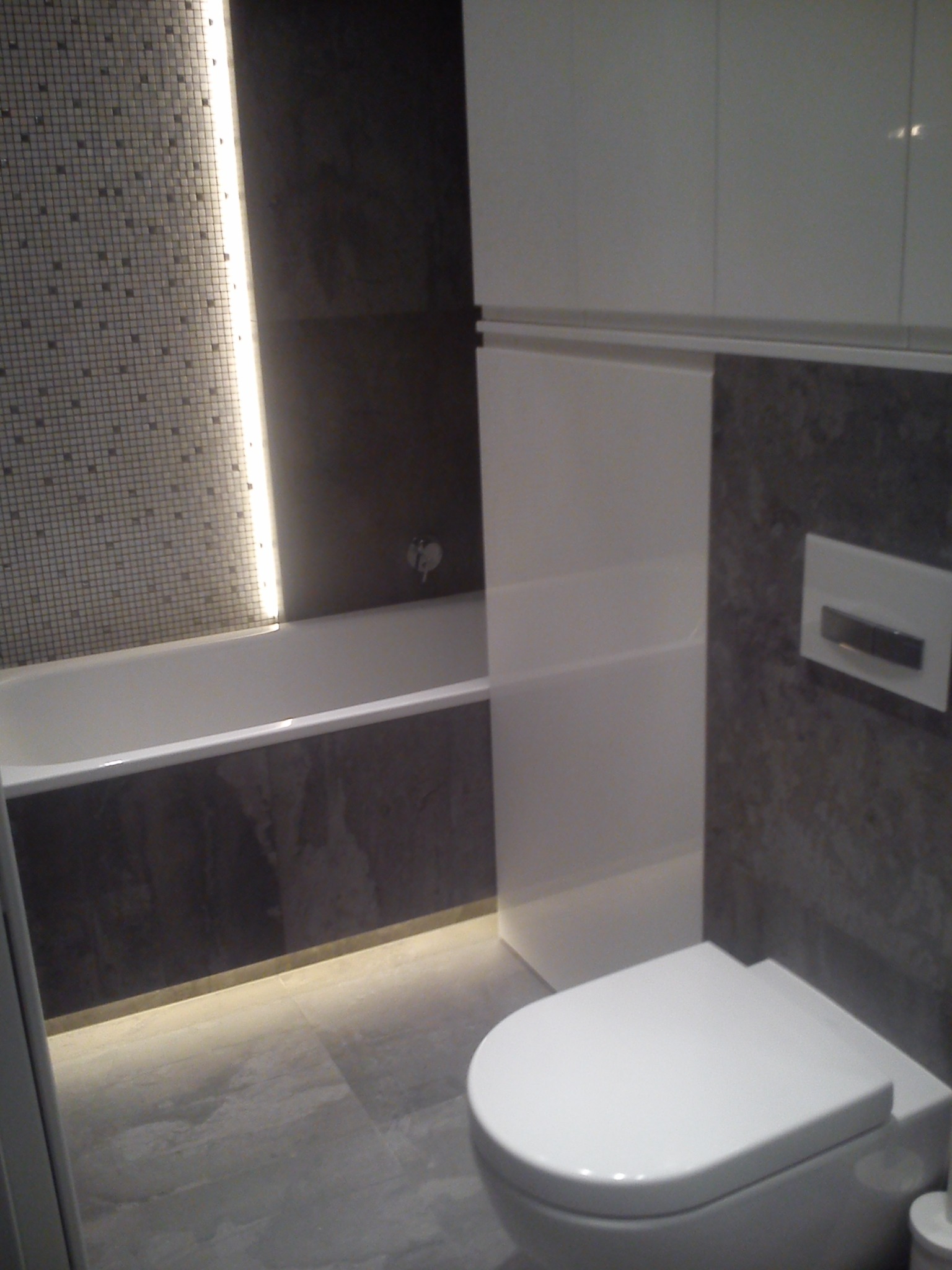 Nowoczesna łazienka z mozaikową ścianą nad wanną, podświetleniem LED, szarą płytką imitującą kamień i wiszącą toaletą.