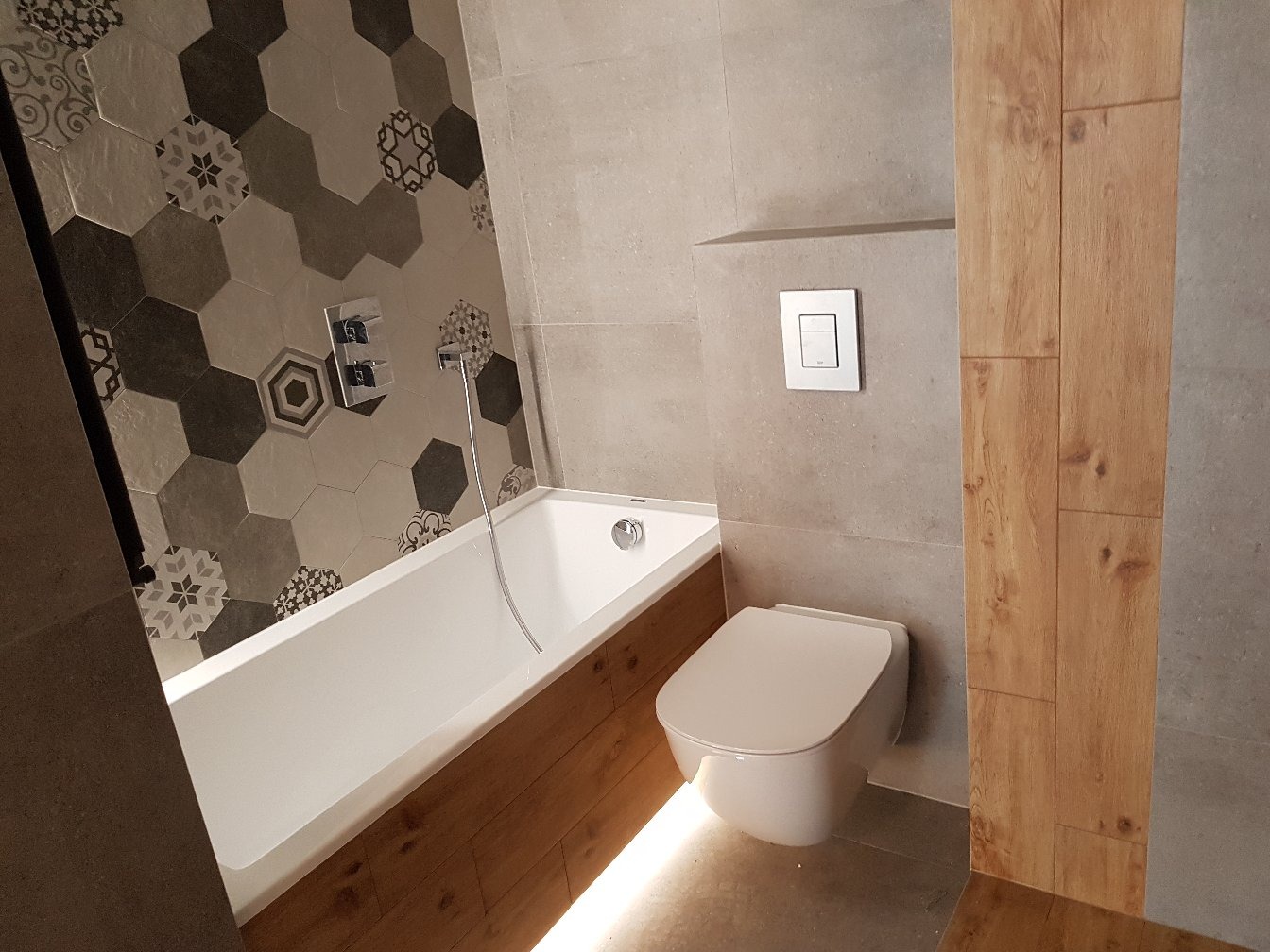 Nowoczesna łazienka z heksagonalnymi płytkami w odcieniach szarości i beżu, drewnopodobną obudową wanny z podświetleniem LED, wiszącą miską WC i armaturą w kolorze chromu.