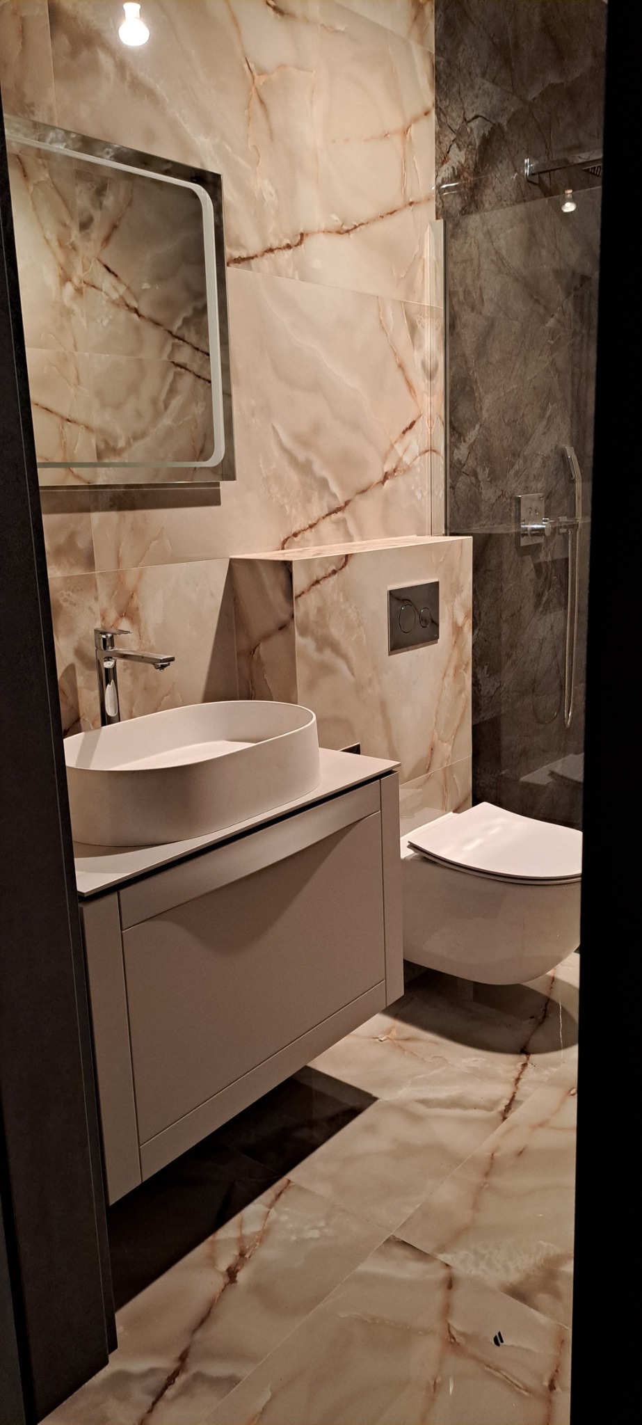 Wykończona łazienka z beżowymi płytkami imitującymi marmur na ścianach i podłodze, owalna umywalka nablatowa na szarej szafce, wisząca toaleta i szklana kabina prysznicowa z ciemnymi płytkami.