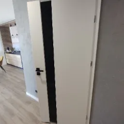 Nowoczesne, białe drzwi wewnętrzne z czarnym, pionowym akcentem, zamontowane w mieszkaniu o szarych ścianach i drewnianej podłodze, widok na kuchnię w tle.