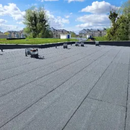 Szary, płaski dach pokryty papą termozgrzewalną z elementami wentylacyjnymi i zielenią w tle. Widoczne budynki mieszkalne i błękitne niebo.